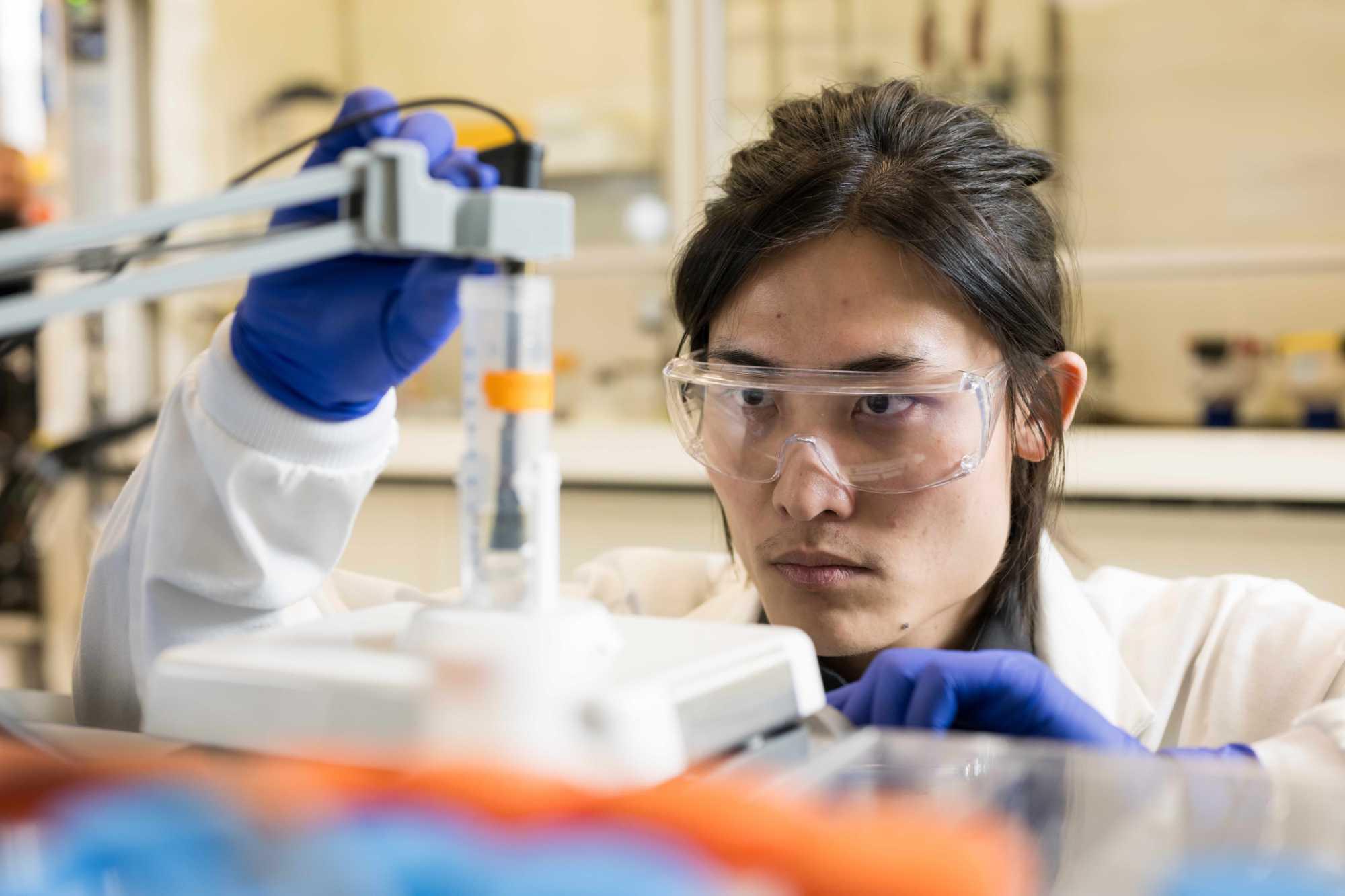 A scientist wearing lab googles and gloves examines a beaker of water containing PFAS chemicals.