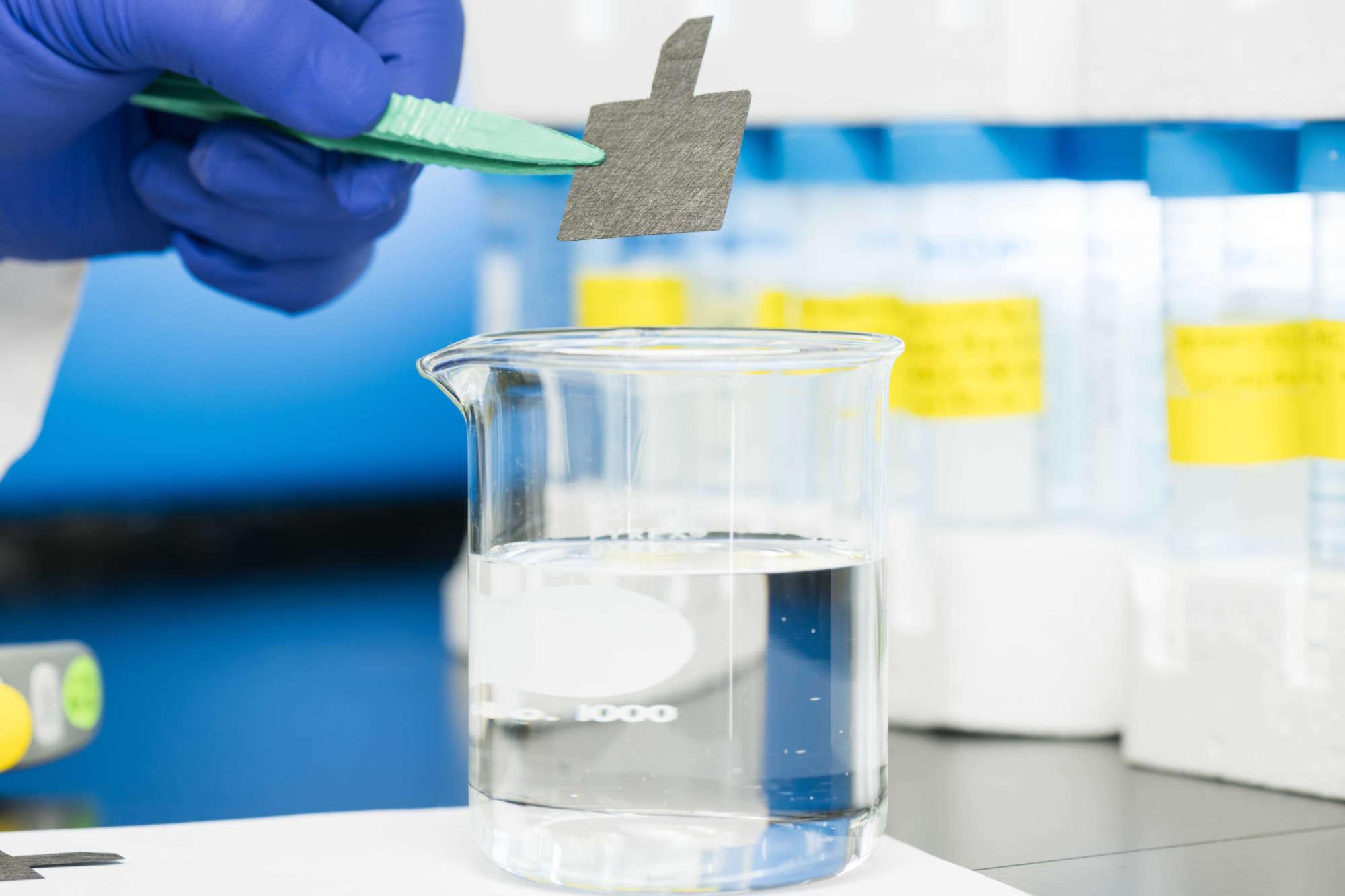 A gloved hands uses tweezers to hold carbon paper above a glass beaker filled with water to illustrate a new technique to remediate PFAS chemicals.